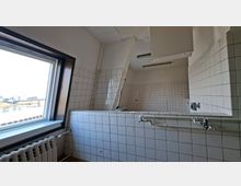 Innenraum einer ehemaligen Großküche oder eines Arbeitsraums mit weiß gefliesten Wänden, großen Abluftkästen unter der Decke und sichtbaren Rohrleitungen über einer gefliesten Trennwand. Links befindet sich ein großes Fenster mit Blick nach draußen und darunter ein Heizkörper.