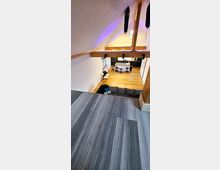 Raum mit offener Balkendecke und moderner Einrichtung, darunter ein Küchenbereich mit einer Insel, Barhockern und schwarzen Schränken auf Holzboden, sowie eine graue Sitzgelegenheit im unteren Bereich. Oben befindet sich ein Podest mit grauem Bodenbelag.