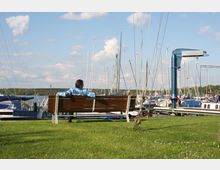 Person auf einer Holzbank am Ufer eines Yachthafens, im Hintergrund liegen Segelboote mit hohen Masten vor Anker; grüne Wiese im Vordergrund und blauer Himmel mit wenigen Wolken.