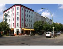 Straßenszene mit einem mehrstöckigen, pastellfarbenen Gebäude mit rosa Akzenten, im Erdgeschoss ein Café mit gelben Markisen. Im Vordergrund eine Kreuzung mit Straßenbahngleisen, geparkten Fahrrädern, Bäumen und einem weißen Transporter.