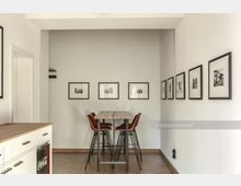 Minimalistisch eingerichteter Raum mit einem hohen Holztisch und vier Barstühlen. An den weißen Wänden hängen mehrere gerahmte Schwarz-Weiß-Fotografien; links befindet sich eine Küchenzeile mit Holzarbeitsplatte.