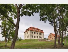 Historisches Herrenhaus mit heller Fassade und rotem Walmdach, umgeben von einer Rasenfläche mit vereinzelten Bäumen im Vordergrund; daneben ein kleineres Gebäude aus Backstein mit ähnlichem Dach.