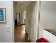 Blick in einen schmalen Flur mit hellem Holzboden und weißen Wänden. Im Hintergrund befindet sich ein Fenster mit weißen Vorhängen, ein großer Spiegel lehnt an der Wand, und links ist ein Wandbild zu sehen.