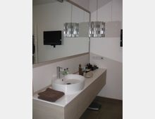 Modernes Badezimmer mit Waschtisch aus Holzoptik, rundem weißen Waschbecken und großem Spiegel. Zwei silberne Hängelampen, ein wandmontierter Flachbildschirm und Accessoires wie Seifenspender und Handtuch ergänzen die Ausstattung.