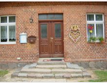 Eingangsbereich eines Gebäudes mit rotem Backstein. Zentral stehen hölzerne zweiflügelige Türen, flankiert von zwei Fenstern mit weißen Rahmen und Gardinen. Links neben der Tür befinden sich ein Briefkasten und ein Infokasten, rechts ein dekoratives Schild mit der Aufschrift „Kita Kinderträume“ und ein bepflanzter Blumenkasten.