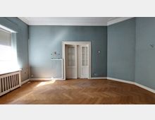 Leerer Raum mit blauen Wänden und einem Fischgrätenparkettboden. Im Raum befinden sich ein großes Fenster mit Jalousien auf der linken Seite, sowie eine zweiflügelige weiße Tür mit Glaseinsätzen an der gegenüberliegenden Wand.