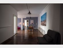 Wohnzimmer mit Holzboden und minimalistischer Einrichtung. Im Vordergrund steht ein graues Sofa, im Hintergrund ein Holztisch mit Stühlen, an der Wand ein großes Landschaftsfoto und dekorative Lampen.