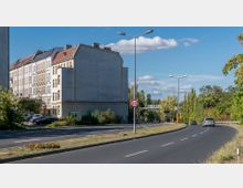 Straßenszene mit Häuserfassaden auf der linken Seite, einem großen, unbemalten Giebel und einer Hauptstraße mit Fahrbahnteilung rechts. Die Umgebung enthält begrünte Bereiche und Bäume, ein Tempo-60-Verkehrsschild und vereinzelte Autos auf der Straße.