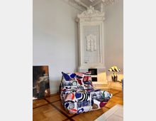 Ein stilvoll eingerichtetes Zimmer mit einer verzierten weißen Kachelheizung im klassischen Design, einem bunten Sessel mit abstraktem Muster, und einer gestreiften Tischlampe auf einem Glastisch. Der Boden besteht aus einem Holzparkett mit geometrischem Muster.