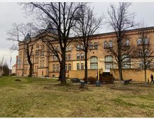 Backstein-Gebäude im neoromanischen Stil mit hohen, schmalen Fenstern und Türmchen, umgeben von einer Grünfläche mit Bäumen und Bänken. Im Vordergrund ist eine gepflegte Wiese mit einzelnen Wegen sichtbar, während der Himmel bewölkt ist.