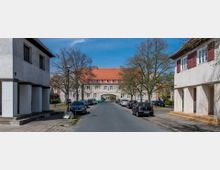 Straßenszene in einem Wohngebiet mit symmetrischen Häusern auf beiden Seiten, die cremefarbene Fassaden und rote Dächer haben. Im Hintergrund befindet sich ein großes Gebäude mit einem Torbogen, flankiert von Bäumen, während Autos am Straßenrand parken.