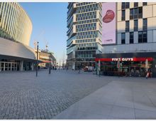 Städtischer Platz mit moderner Architektur, flankiert von einem abgerundeten Glasgebäude links und einem Hochhaus mit geometrischen Glasfassaden rechts. Im Vordergrund liegt ein Restaurant der Kette "Five Guys", während im Hintergrund Baukräne und weitere Gebäude sichtbar sind.