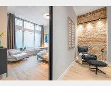 Modernes Apartmentzimmer mit großen Fenstern und grauen Vorhängen, hellem Holzboden sowie Bett und kleinem Sofa. Rechts daneben eine Sitzecke mit schwarzem Lounge-Sessel, Beistelltisch und freiliegender Ziegelwand mit Schienenbeleuchtung.