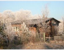 Kleines Backsteinhaus mit Holzanbau und Schrägdach, umgeben von winterlicher Landschaft mit frostbedeckten Bäumen und Sträuchern. Ein Maschendrahtzaun grenzt das Grundstück ein.