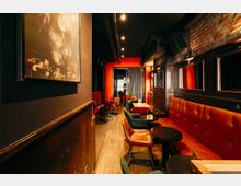 Schmaler Bar- oder Loungebereich mit roten gepolsterten Sitzbänken, kleinen runden Tischen und gemischten Stühlen entlang eines Holzfußbodens. Dunkle Decke mit sichtbaren Rohrleitungen und Spotbeleuchtung, rechts eine freiliegende Ziegelwand mit Spiegeln und am Ende ein rot gerahmter Durchgang mit Vorhang.
