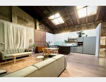 Offener Loftraum mit hoher Decke, sichtbaren Holzbalken und großen Oberlichtern sowie freigelegten Backsteinwänden. Im Vordergrund ein Wohnbereich mit Sofas und Couchtisch, daneben ein Esstisch und eine moderne Küchenzeile mit Regalen und Pflanzen.
