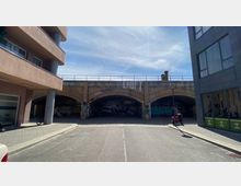 Straßenansicht eines Backsteinbogens unter einer Eisenbahnbrücke, verziert mit Graffiti und dem Schriftzug "LÄUFT". Links und rechts stehen moderne Gebäude, vorne befindet sich eine gepflasterte Straße mit einem geparkten Roller.