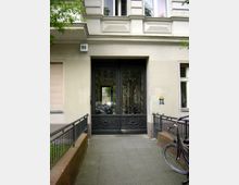 Eingangsbereich eines Wohnhauses mit einer kunstvoll verzierten, schwarzen Doppeltür aus Metall. Die Fassade ist beige, es gibt ein schmiedeeisernes Geländer auf beiden Seiten des Zugangswegs und ein angebundenes Fahrrad steht rechts im Bild.
