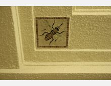 Mosaikfliese mit der Abbildung einer stilisierten Biene, eingebettet in eine gerahmte, strukturierte Wand mit gelblicher Beleuchtung. Die Fliese ist von einem roten Rand umgeben.