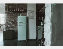 Küche mit Backsteinwand, einem mintgrünen Retro-Kühlschrank, Herd und Küchenutensilien. Im Vordergrund ist eine helle Säule mit dekorativem Poster, links ein Fenster mit Tageslicht.