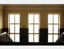 Treppenhaus mit drei großen rechteckigen Fenstern, die viel Tageslicht hereinlassen. Die Wände sind hell, und die Treppengeländer sowie die Verkleidungen der unteren Wandbereiche bestehen aus dunklem Holz.