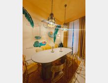 Ein Raum mit einem großen ovalen Tisch aus hellem Material, umgeben von transparenten Plastikstühlen. An der Wand befindet sich eine farbenfrohe Wandmalerei mit tropischen Motiven, daneben hängen drei verzierte Pendelleuchten von der Decke.