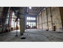 Verlassene Industriehalle mit hohen Backsteinwänden, großen Fenstern und teilweise zerstörtem Boden. Im Raum sind Überreste von Leitungen, Metallgerüsten und alten Geräten erkennbar.