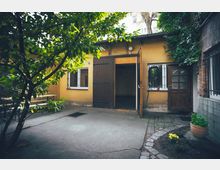 Kleiner Hinterhof mit gelb gestrichener Fassade, zwei Fenstern mit Gitter, einer offenen Holztür und einem Holztor. Im Vordergrund befinden sich ein Baum mit grünen Blättern, ein gepflasterter Weg, eine Blumentopfpflanze und eine gelbe Blume.