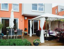 Moderne Terrasse eines Hauses mit orangefarbener Fassade, ausgestattet mit Holzboden, einer Sitzgruppe unter einem weißen Pavillon mit Vorhängen und einem Esstisch mit schwarzen Stühlen. Im Vordergrund befinden sich Pflanzen, und im Hintergrund ist ein Sichtschutzzaun aus Bambus sichtbar.