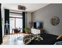 Modernes Wohnzimmer und Schlafzimmer in einem Raum mit hellen Holzböden. Links ein Schreibtisch vor einer großen Fensterfront mit dunklen Vorhängen, rechts eine graue Wand mit einem Fernseher auf einem weißen Sideboard und goldenem Wanddekor. Im Vordergrund ein Bett mit dunkler Bettdecke und einer Kunstfelldecke.