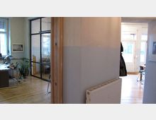 Büroinnenraum mit Holzboden, einer Wand in hellen Farben und einer Glas- sowie Metalltrennwand, die zwei Bereiche abteilt. Links steht ein Schreibtisch mit Bürostuhl und einer Pflanze, rechts führt ein Türbereich zu einem anderen Raum mit einem Glasfenster und einer Außentür.