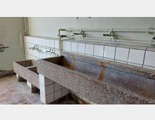 Waschraum mit großen, rechteckigen Waschbecken aus grobem Beton, gefliestem Hintergrund mit weißen Keramikfliesen und sichtbaren Rohrleitungen an der Wand. Der Raum zeigt Anzeichen von Abnutzung und hat einen Boden aus rötlich-beigen Fliesen.