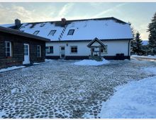 Schneebedeckter Hof mit Kopfsteinpflaster, eingerahmt von einem weißen Einfamilienhaus mit dunklem Dach und einem Holzgebäude. Im Vordergrund steht ein überdachter Brunnen, und im Hintergrund sind Bäume sichtbar.
