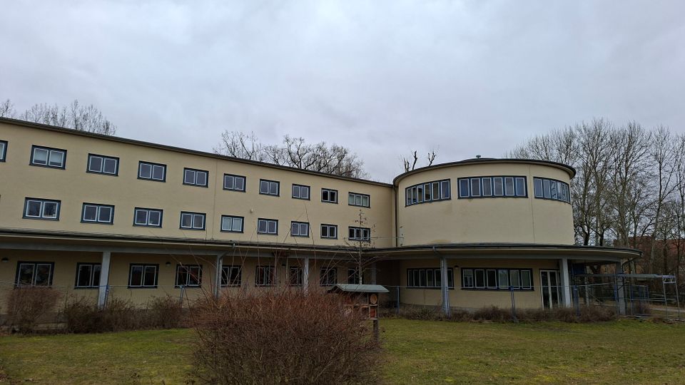 Beiges, dreistöckiges Gebäude mit vielen rechteckigen Fenstern und einem markanten runden Anbau an der rechten Seite. Davor liegen eine Rasenfläche mit niedrigen Sträuchern und ein überdachter Eingangsbereich; im Hintergrund stehen laublose Bäume unter bewölktem Himmel.