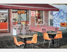 Außenbereich eines kleinen Restaurants mit Backsteinfassade. Vor dem Eingang stehen orangefarbene Plastikstühle, Tische sowie lange Bänke mit rot-weiß karierten Tischdecken; im Hintergrund ist ein Wandgemälde mit Bergmotiven zu sehen.