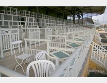 Reihen von Tribünenplätzen mit weißem Geländer und vereinzelten weißen Stühlen, in einer überdachten Struktur mit Metallträgern. Im Hintergrund sind weitere Tribünenbereiche und Gebäude sichtbar.