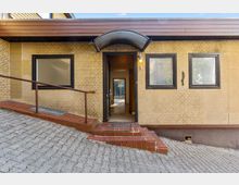 Eingangsbereich eines Backsteingebäudes mit offener Tür, kleinem gewölbtem Vordach und drei dunkel gerahmten Fenstern. Vor dem Eingang führen eine flache Rampe mit Geländer und niedrige Stufen über einen gepflasterten, leicht abfallenden Hof.