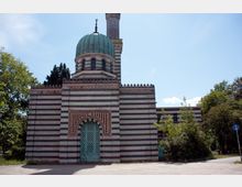 Historisches Gebäude mit einer Kuppel und einem Minarett, gestaltet aus horizontal gestreiften, bunten Ziegelsteinen. Der Eingangsbereich ist mit einem verzierten Torbogen und einem grün gestrichenen Gittertor versehen, umgeben von vereinzelten Bäumen und Pflanzen.
