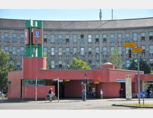 Eingangsbereich einer U-Bahn-Station mit einem roten, flachen Gebäude und einem Turm, der ein U-Bahn-Symbol trägt. Im Hintergrund steht ein mehrstöckiges Gebäude mit grauer Fassade und vielen Fenstern; Straßenschilder und Fußgänger befinden sich im Vordergrund.