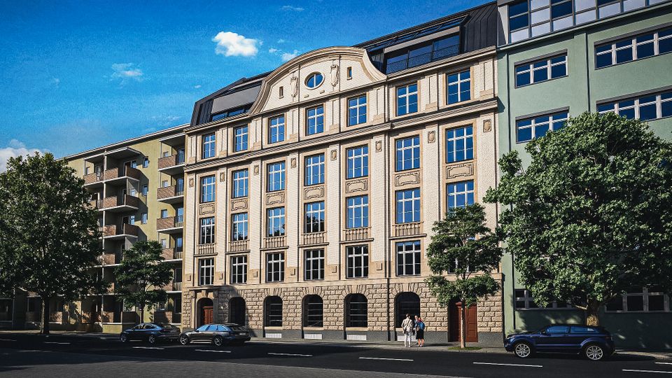 Mehrstöckiges städtisches Gebäude mit klassischer Sandsteinfassade, symmetrischen Fensterreihen und rundbogigen Öffnungen im Erdgeschoss, darüber ein Dachaufbau mit Gauben. Daneben angrenzen moderne Wohngebäude mit Balkonen; davor verläuft eine breite Straße mit parkenden Autos und Bäumen am Gehweg.