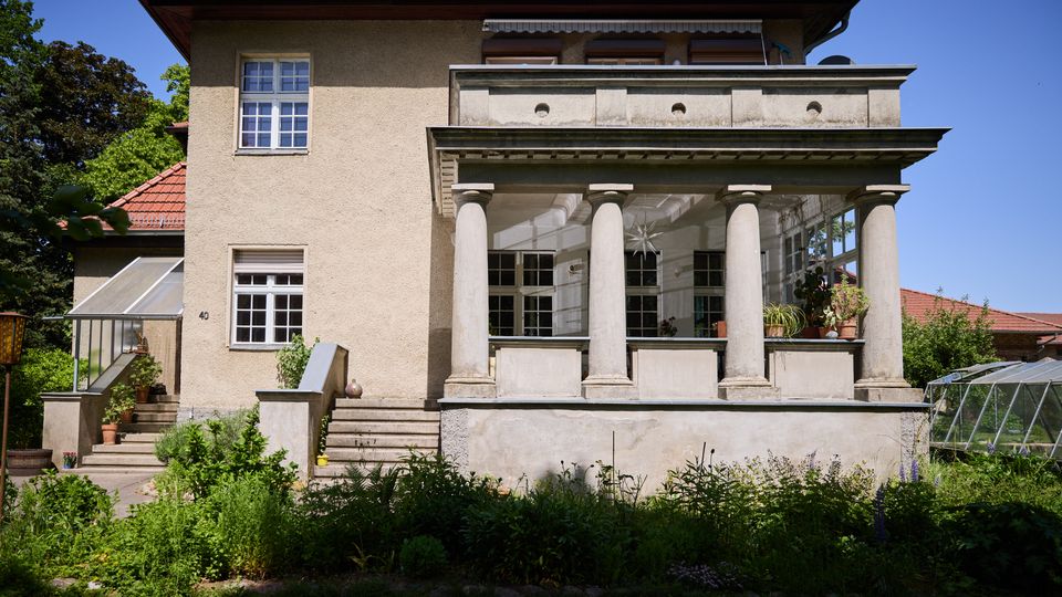 Zweistöckiges Wohnhaus mit Putzfassade und rotem Ziegeldach, davor eine erhöhte Veranda mit vier Säulen, großer Balkonbrüstung und breiten Treppenstufen. Im Vordergrund liegt ein dicht bepflanzter Garten; seitlich sind ein kleines Gewächshaus und weitere Gartenbereiche zu sehen.