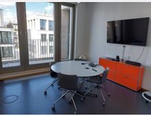 Besprechungsraum mit modernem Design, bestehend aus einem runden Tisch mit vier Bürostühlen, einem großen Flachbildschirm an der Wand und einem orangefarbenen Sideboard. Große Fenster geben den Blick auf ein modernes Gebäude im Außenbereich frei.
