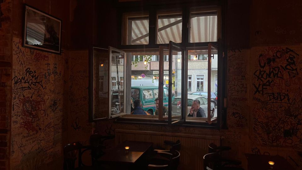 Innenraum eines kleinen Cafés mit dicht beschmierten, graffitiartigen Wänden und dunklen Holztischen mit Stühlen. Ein großes, teilweise geöffnetes Fenster mit Oberlichtern zeigt den Blick auf die Straße mit geparkten Autos und gegenüberliegenden Gebäuden.