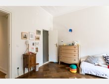 Helles Kinderzimmer mit Holzboden, einer weißen Wand und einer geöffneten Tür. Links steht ein kleiner Holztisch mit einer Lampe, daneben hängen einige gerahmte Bilder; rechts befinden sich eine Kommode mit Spielzeug und ein Sofa mit Kissen.