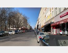 Straße in einer Stadt mit geparkten Autos auf beiden Seiten, flankiert von mehrstöckigen Gebäuden mit Restaurants und Geschäften im Erdgeschoss; kahle Bäume säumen die linke Seite der Straße, und der Himmel ist klar und blau.