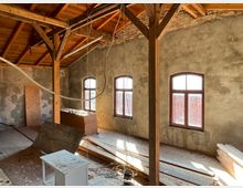 Eine Baustelle in einem Altbau mit unverputzten Wänden, Holzstützbalken und einem Dach aus Holz. Drei große Fenster mit Rundbögen lassen Tageslicht herein, und Baumaterialien liegen im Raum verstreut.