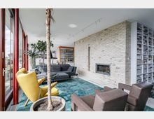 Modernes Wohnzimmer mit einer großen Fensterfront, die viel Tageslicht hereinlässt, einer Backsteinwand mit eingebautem Kamin, einer grauen Eckcouch, gelbem Sessel und Ledersesseln. Ein großer Teppich in Türkis bedeckt den Boden, und Bücherregale sowie Zimmerpflanzen vervollständigen das Ambiente.