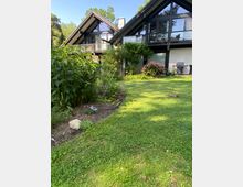 Zwei moderne Häuser mit spitz zulaufenden Dächern aus Glas und Holz, umgeben von einem gepflegten Garten mit grüner Rasenfläche, Blumenbeeten und Sträuchern. Im Hintergrund sind Bäume zu sehen, die eine naturnahe Umgebung andeuten.