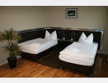 Schlichtes Hotelzimmer mit zwei Einzelbetten, die durch einen Nachttisch mit Leselampen getrennt sind. Der Raum besitzt eine helle Holzfußbodung, eine beige Wand, eine Topfpflanze und ein gerahmtes Bild an der Wand.