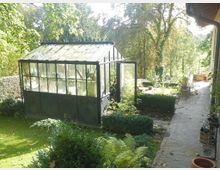 Ein kleines Glashaus mit Metallrahmen und verglasten Wänden steht in einem gepflegten Garten, umgeben von Sträuchern, Farnen und einem gepflasterten Weg, der an einem Gebäude vorbeiführt. Im Hintergrund sind Bäume und eine grüne Landschaft sichtbar.